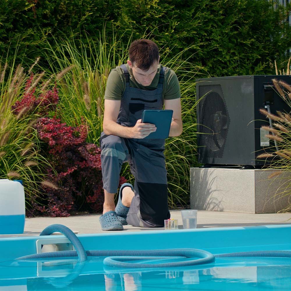 Person crouches by a pool, writing on a tablet beside lush greenery.
