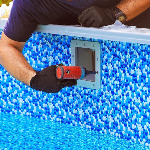 Worker with red drill installing a gray access panel on a blue mosaic-tiled wall