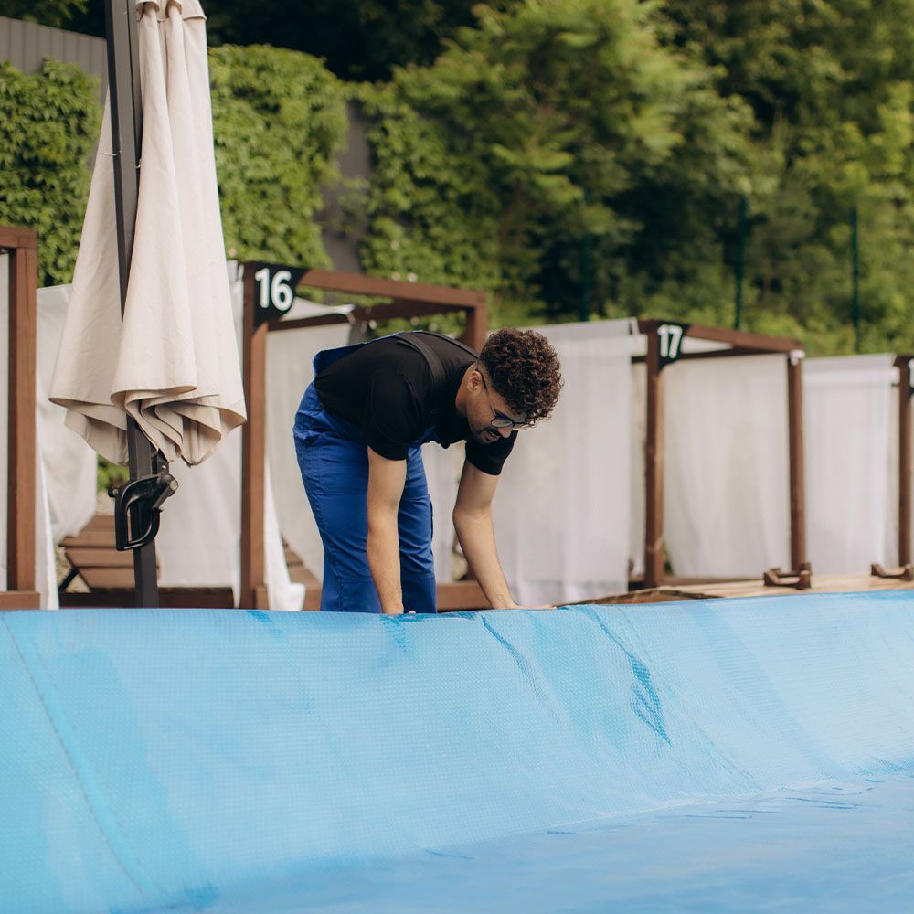 Person in black shirt and blue pants bending over beside a blue pool deck with numbered cabanas in the background