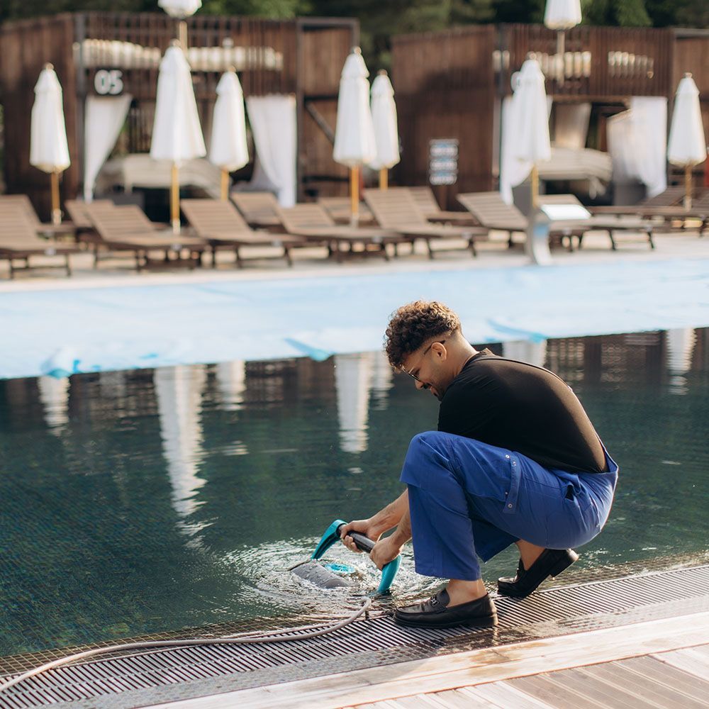 Person crouching by a pool, tying blue swim shoes beside lounge chairs and white umbrellas.