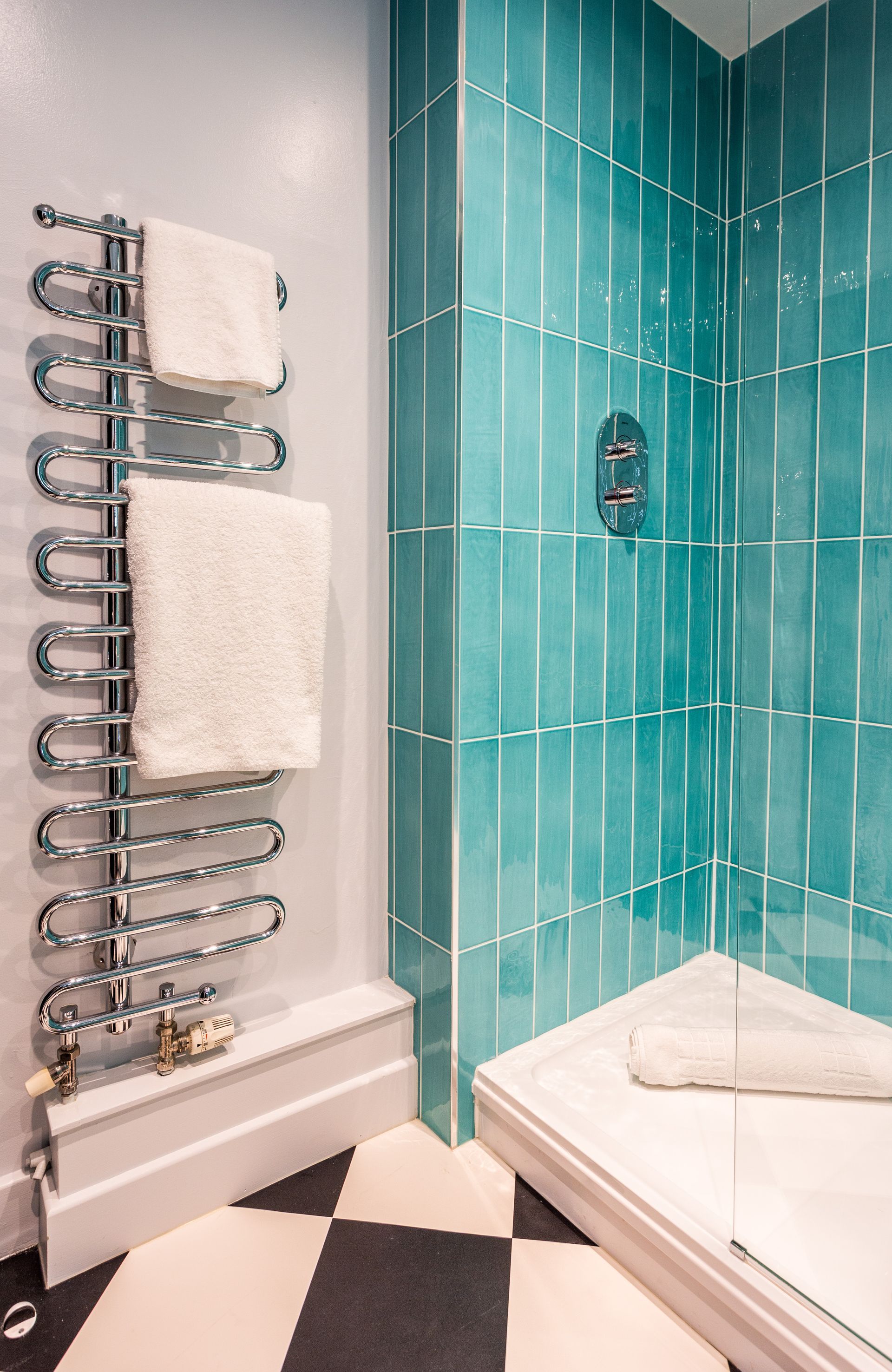 Bathroom with teal tiled shower, chrome towel warmer, and black and white checkered floor.