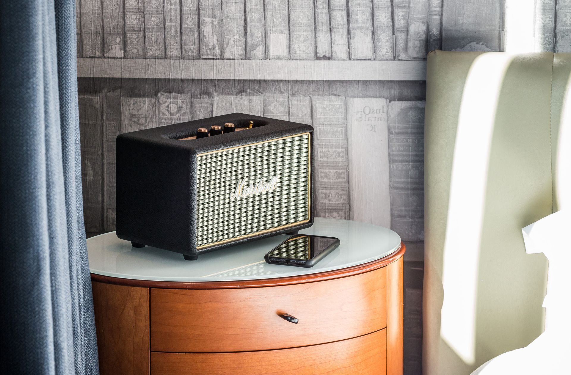 Black Marshall speaker and smartphone on a nightstand, next to a bed, with a bookshelf backdrop.