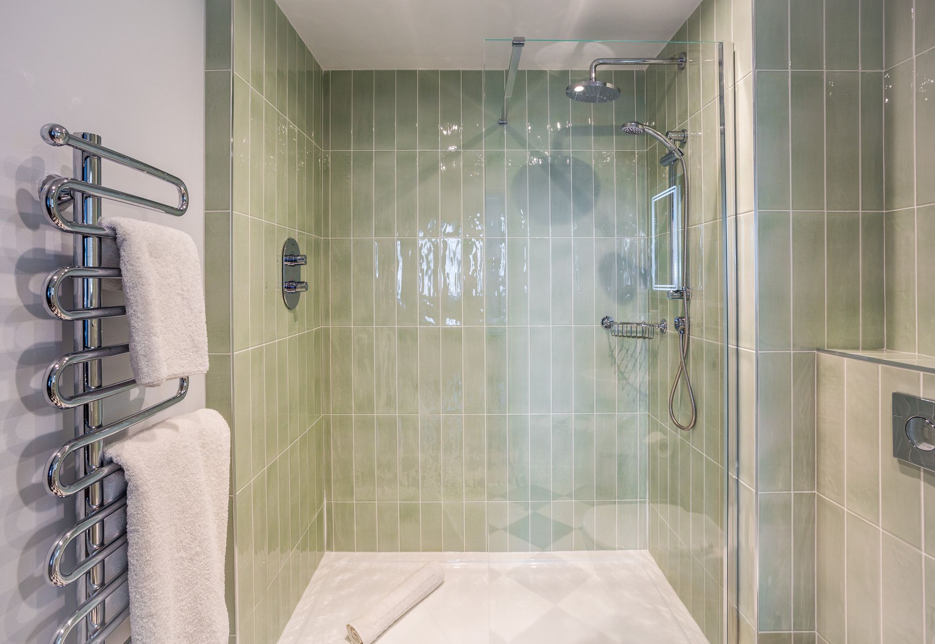 Shower stall with green tiled walls, chrome fixtures, and a towel warmer with towels.