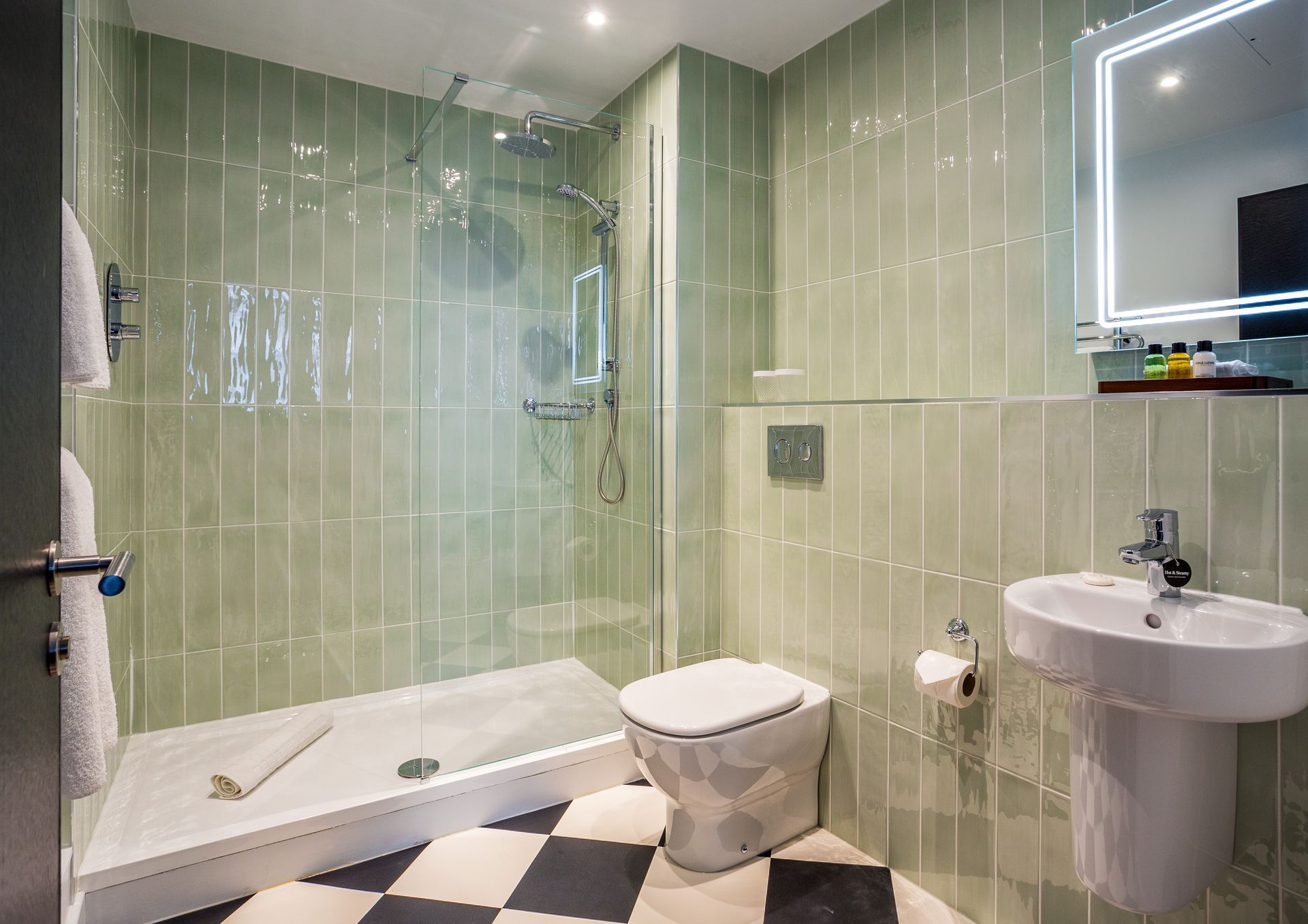 Bathroom with walk-in shower, pedestal sink, toilet, green tiled walls, checkered floor.