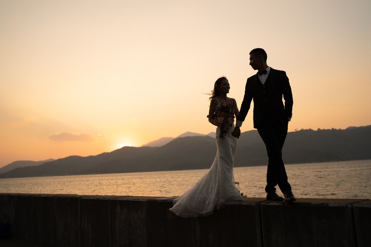 A bride and groom are standing on a pier holding hands at sunset.