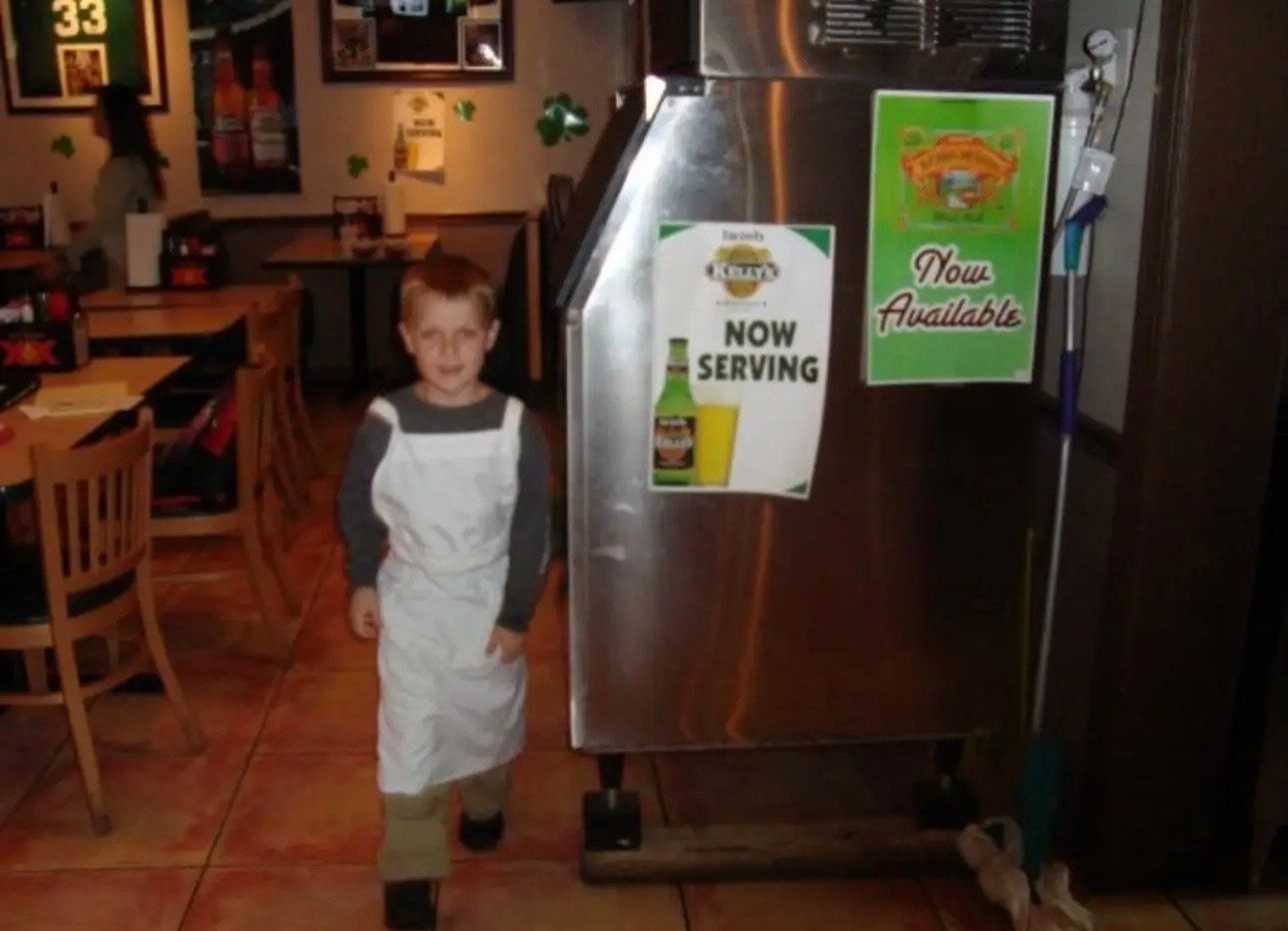 A young boy in an apron stands in front of a now serving sign