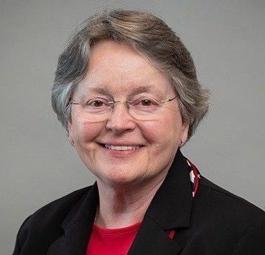 Woman with glasses smiling, wearing a black blazer and red shirt. Gray hair and neutral background.