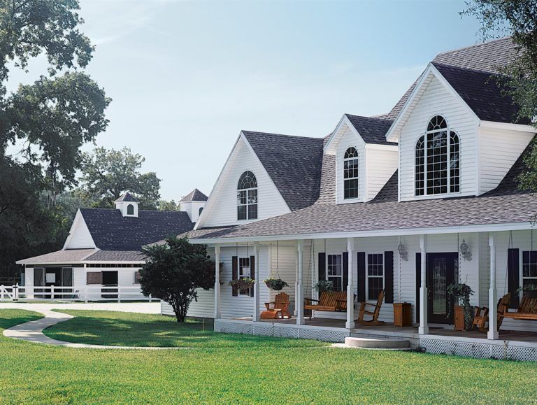 A white farmhouse with a large covered porch and a matching outbuilding on a green lawn under a clear blue sky.
