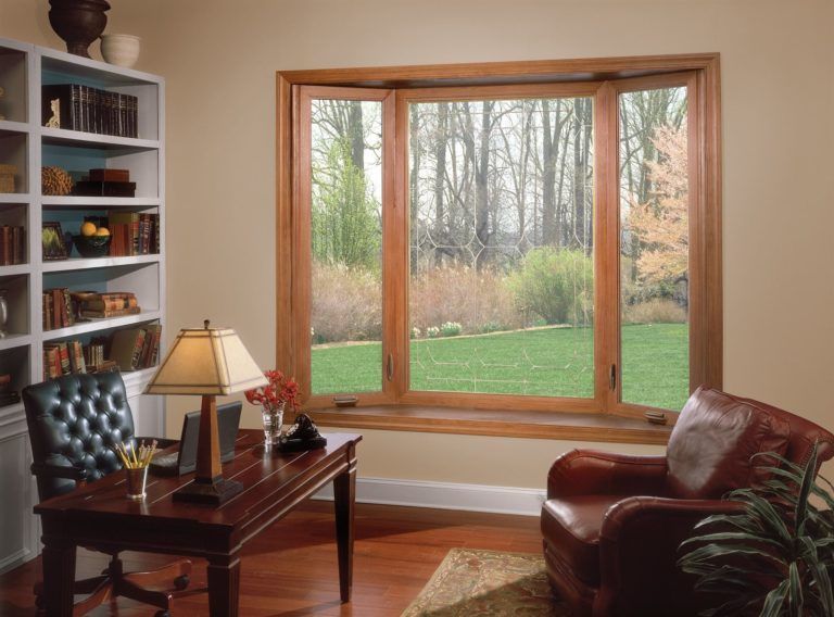 A home office featuring a wooden desk, leather chair, bookshelf, and a large bay window overlooking a garden.
