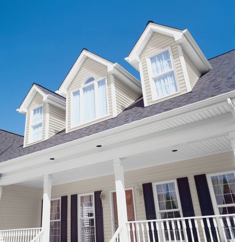 A low-angle view of a beige house with a wrap-around porch, three gabled dormers, and dark shutters under a clear blue sky.
