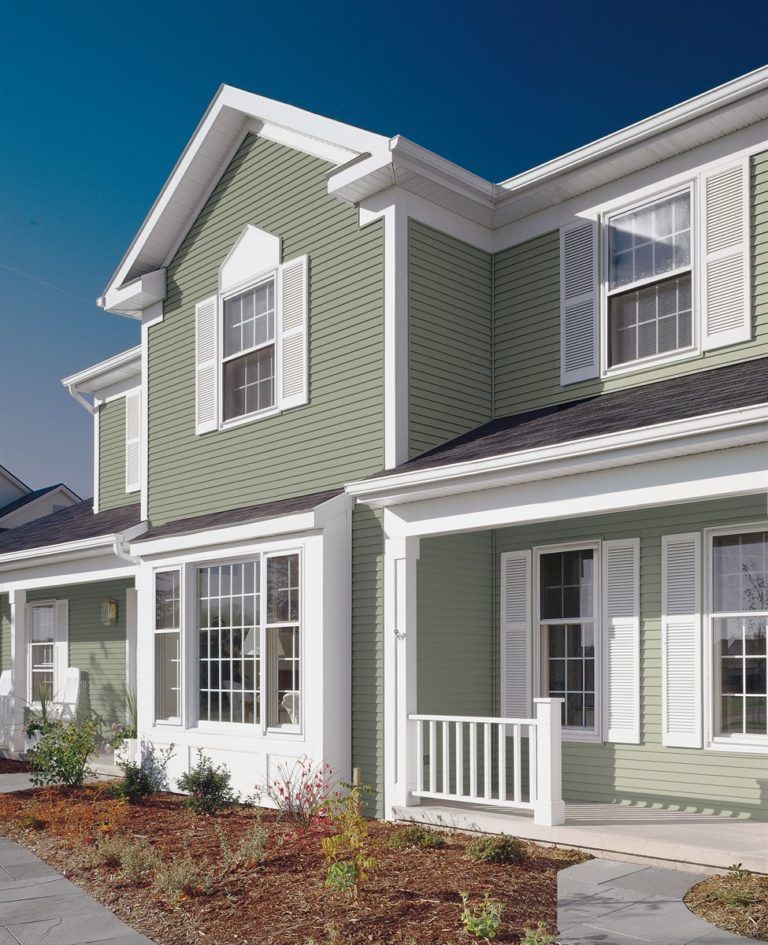 A two-story house with sage green horizontal siding, white trim, white shutters, and a covered porch under a clear sky.