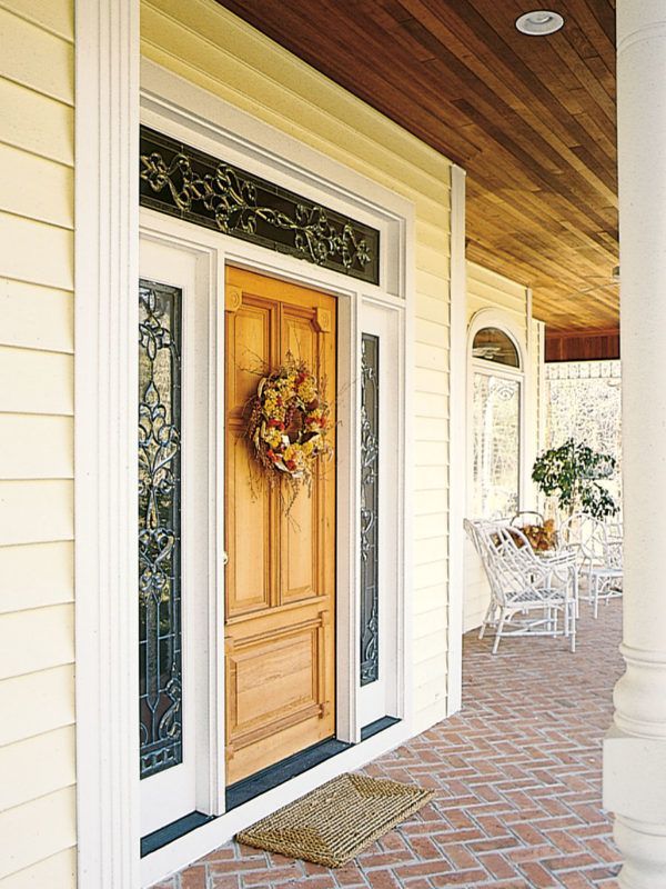 A wooden front door with a wreath, decorative glass panels, and a transom window on a porch with a brick floor.