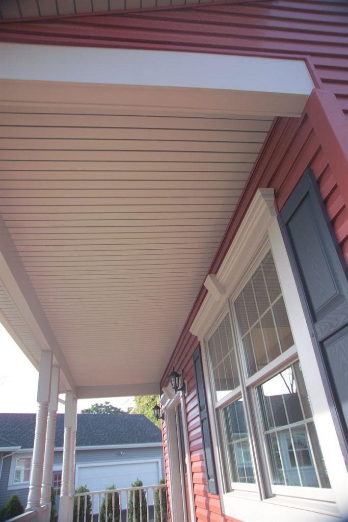 A low-angle view of a red-sided house porch featuring a white ceiling, a window with dark shutters, and white railings.