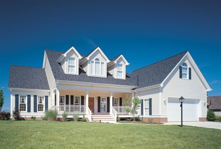A suburban two-story house with white siding, dark shutters, a covered front porch, and three dormers under a blue sky.