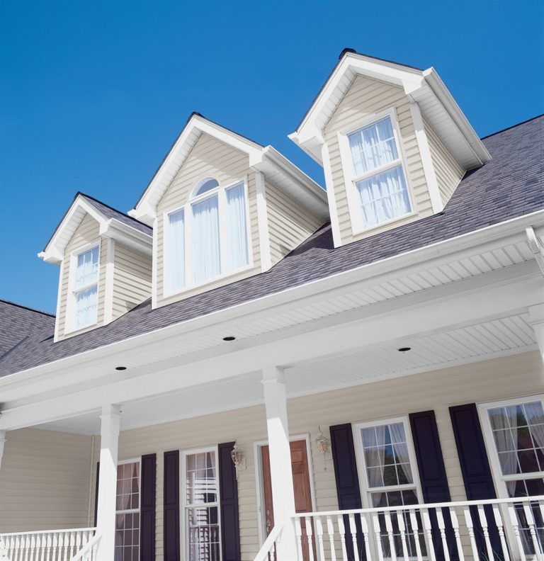 A low-angle view of a beige house with three dormer windows, dark shutters, and a covered front porch against a blue sky.