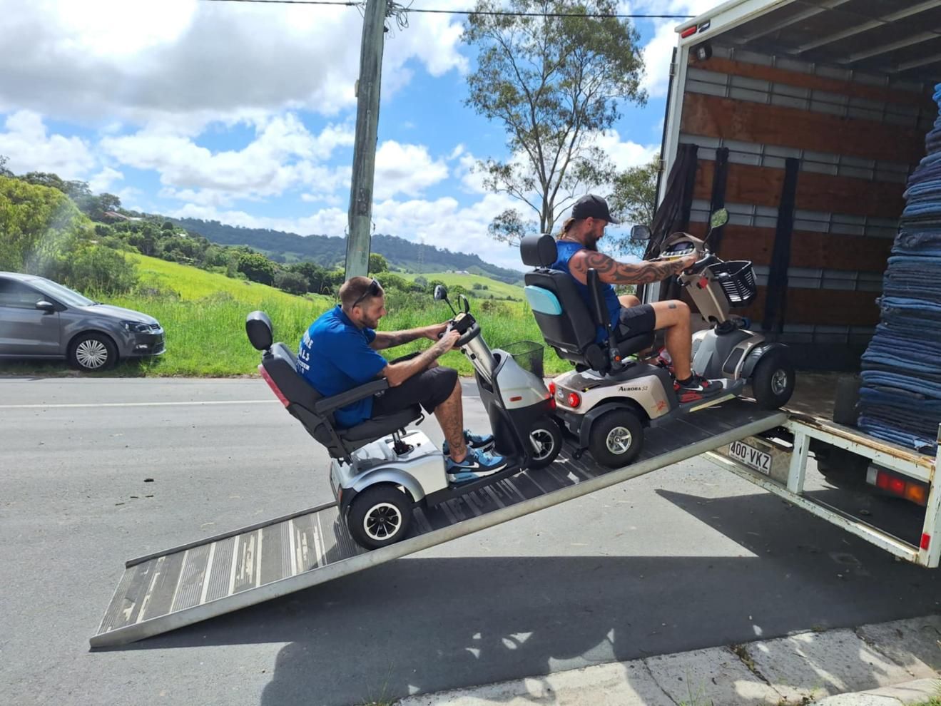 Two Men on Mobility Scooters Driving Up a Ramp Into a Truck on a Road — John's Furniture Removals in Sunshine Coast, QLD