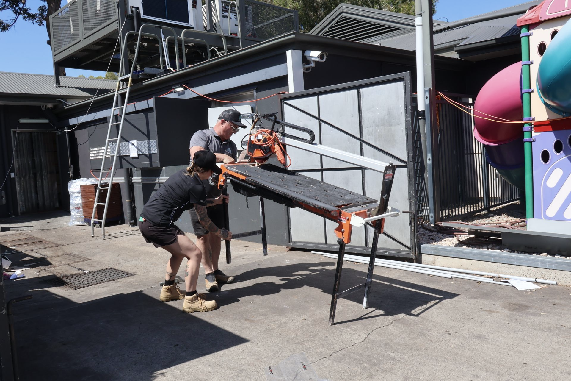 Two People Cutting Wood on A Saw Table Outdoors — Commercial Asset Maintenance In Caloundra, QLD