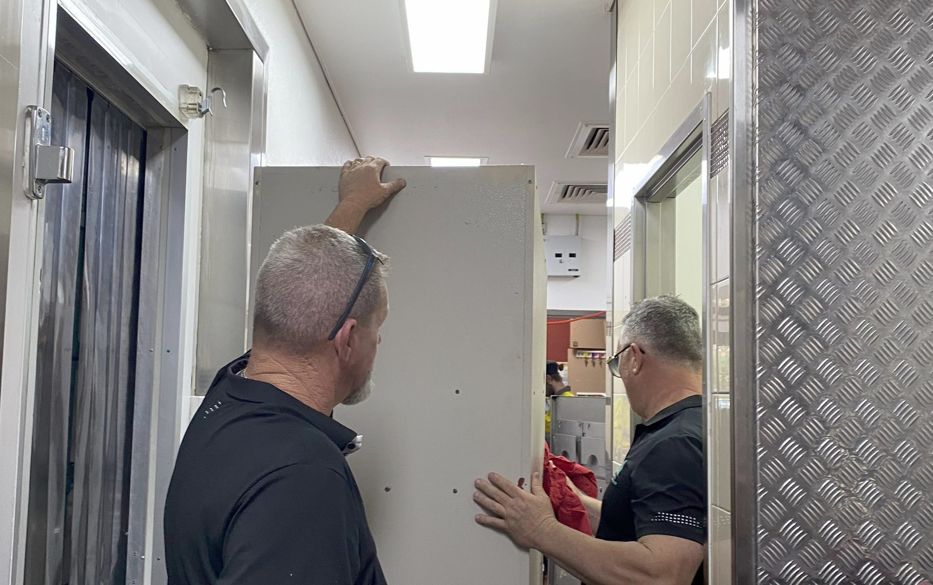 Two Men Install a White Panel in A Narrow Hallway — Commercial Asset Maintenance In Mooloolaba, QLD