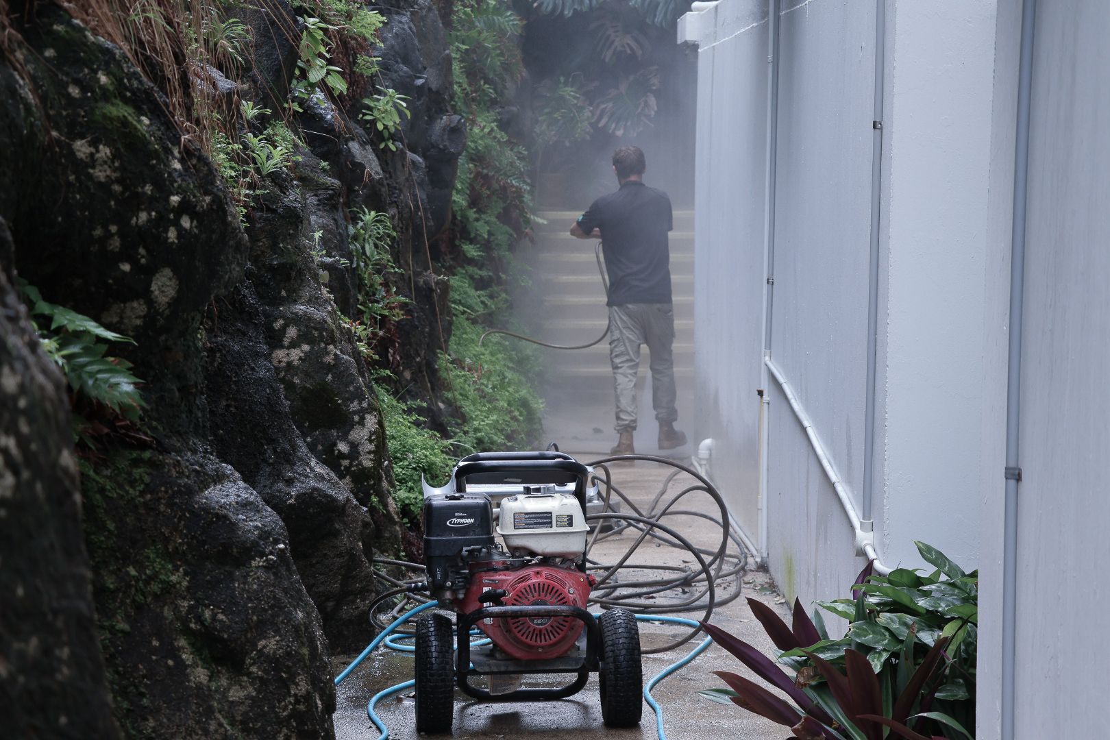 A Man is Pressure Washing a Pathway a Hose in a Garden — Commercial Asset Maintenance In Coolum Beach, QLD