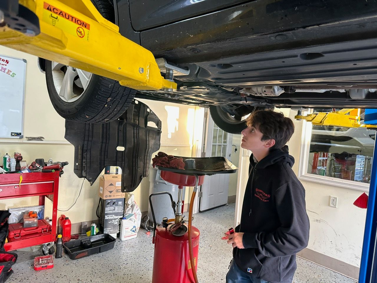 Mechanic checking oil in a car with the hood open in a garage; red toolbox in the background.