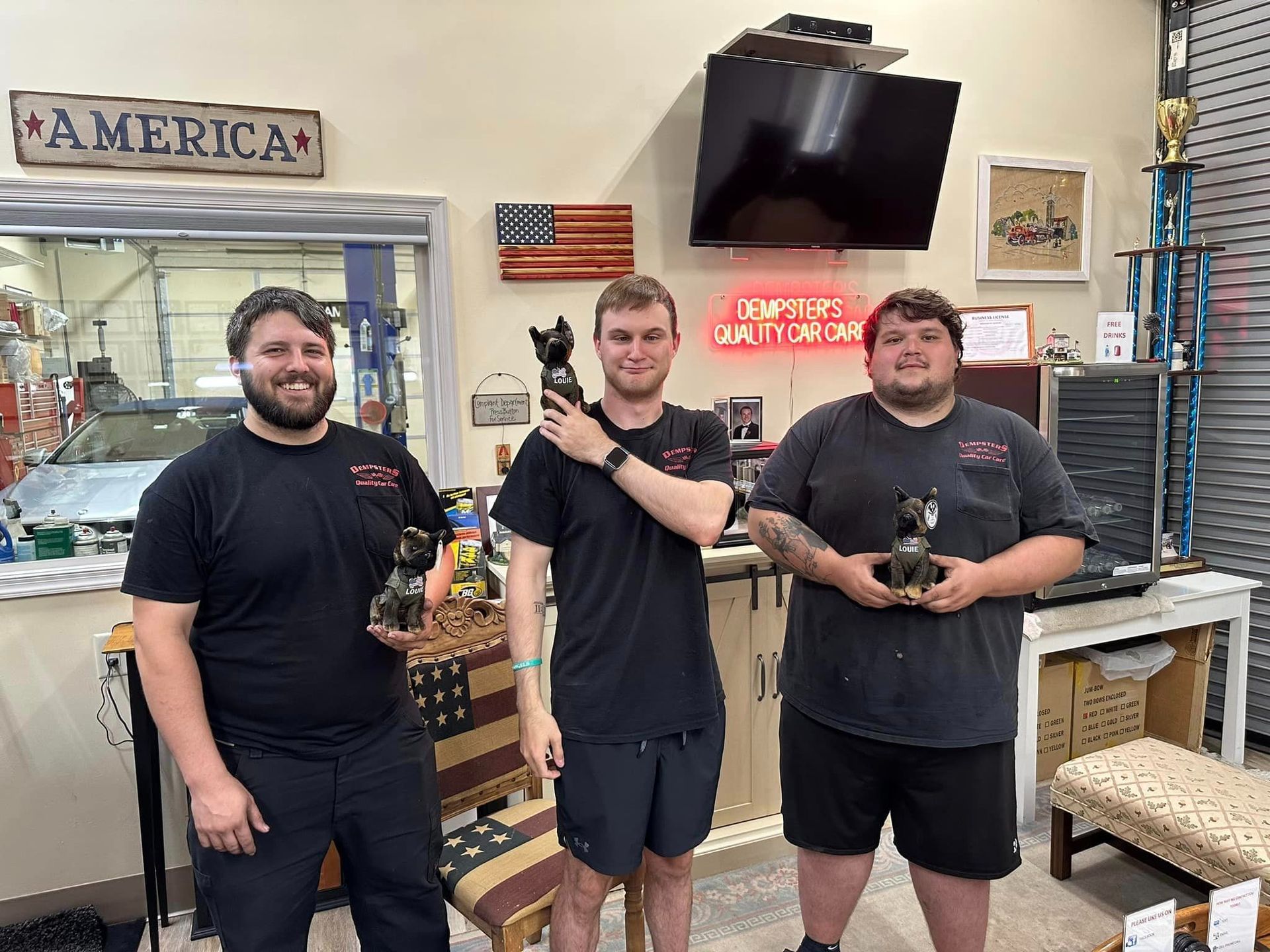 Three men in a shop hold trophies. An American flag and neon sign are in the background.