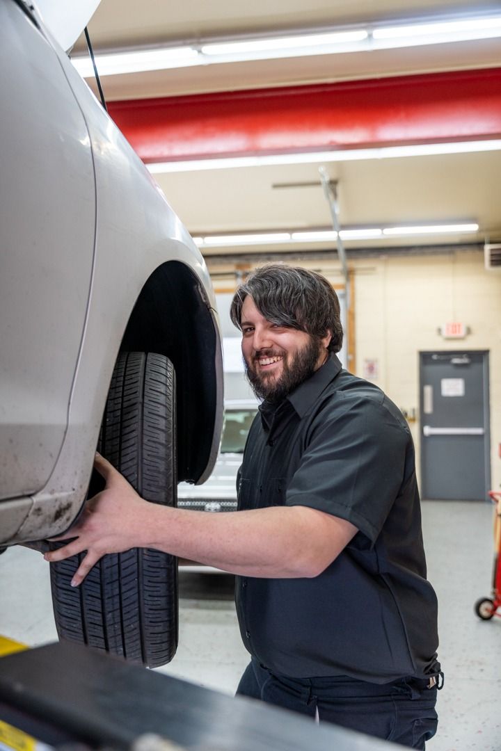 Mechanic in black uniform, inspecting a car tire on a lift, smiling, in a garage setting.