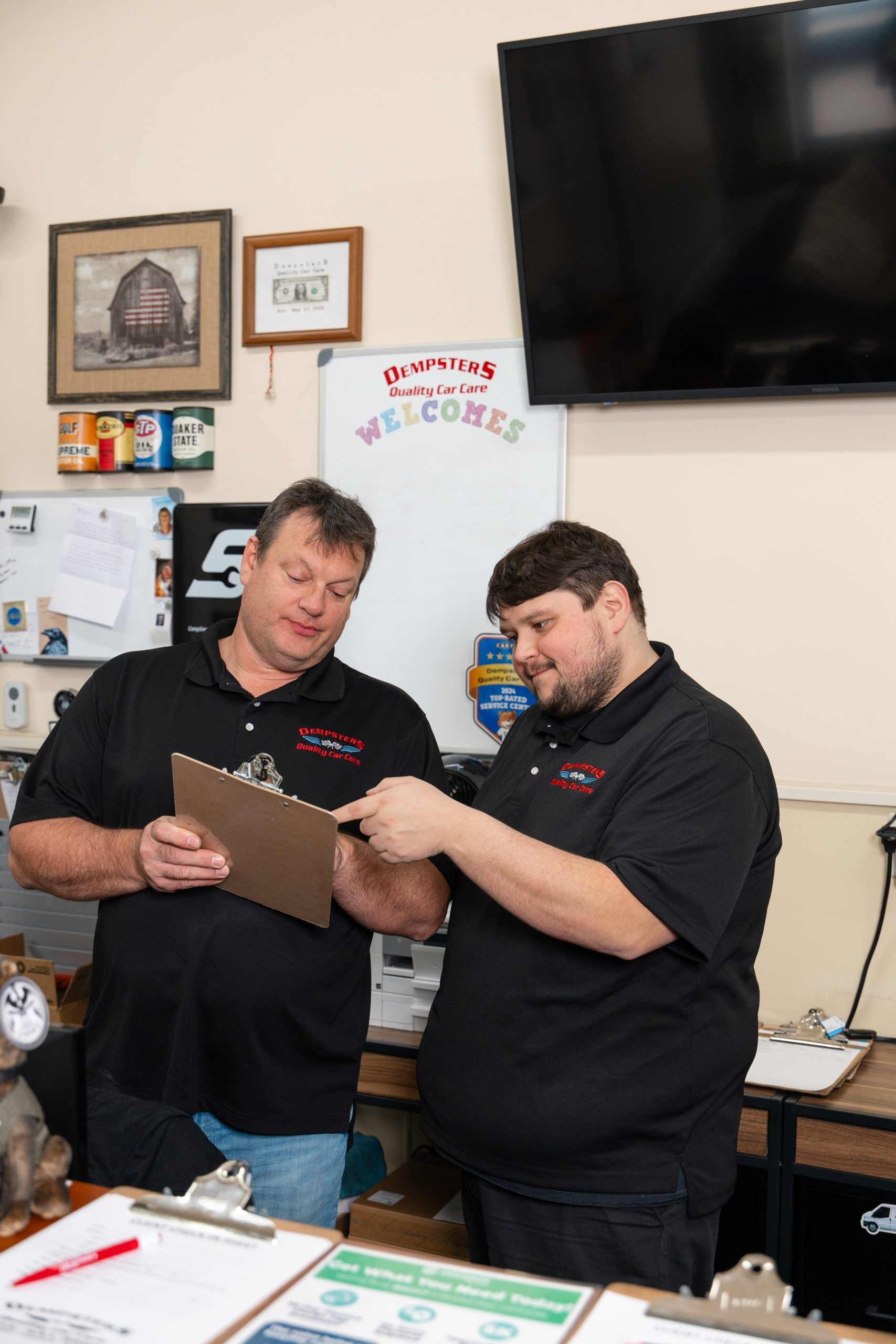 Two men in black shirts review a clipboard in an office, one points.