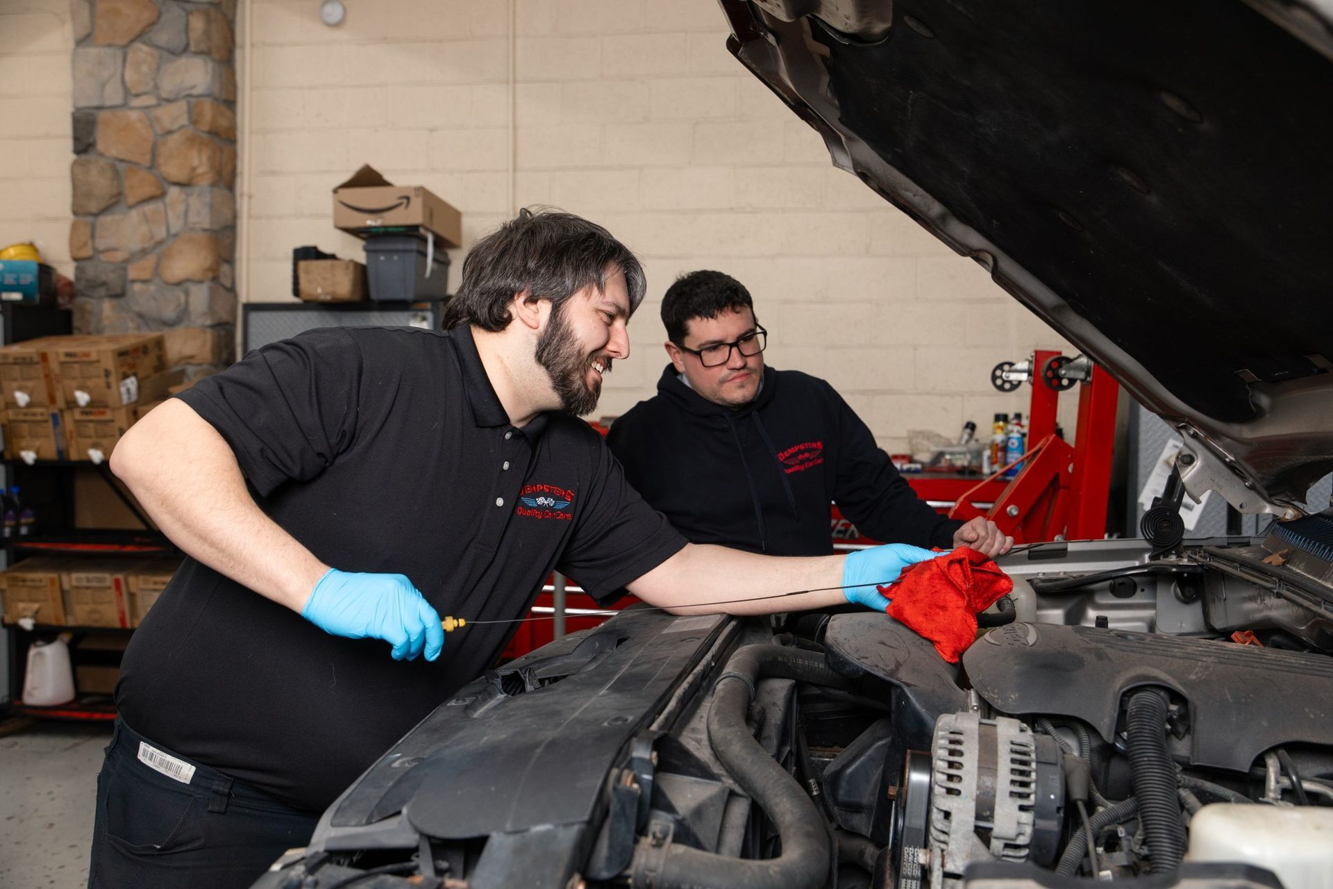 Two mechanics examine a car engine in a garage. One holds a tool, the other a red rag.