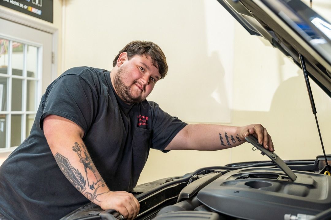 Mechanic leaning over open car hood, inspecting engine, wearing black shirt with tattoos.