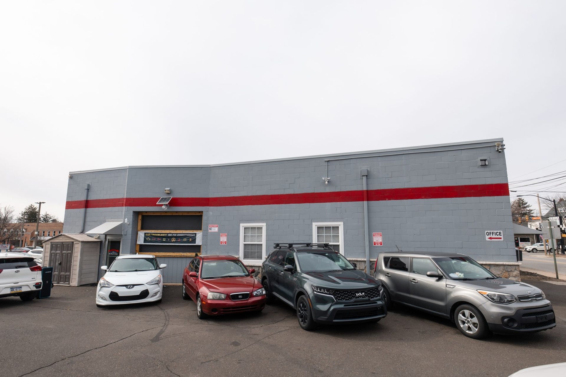 Cars parked in front of a gray building with a red stripe. Cloudy sky.