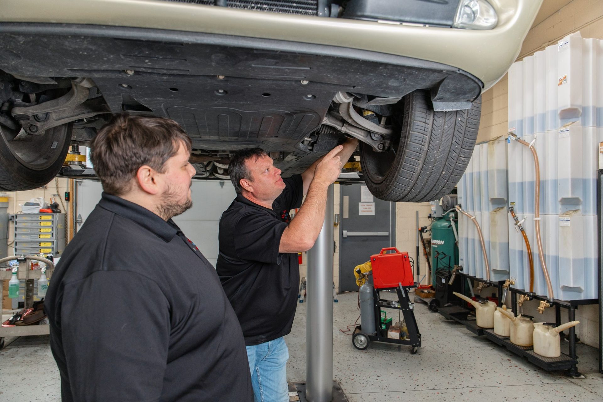 Two mechanics working on a car raised on a lift in a garage. One is under the car, the other observing.