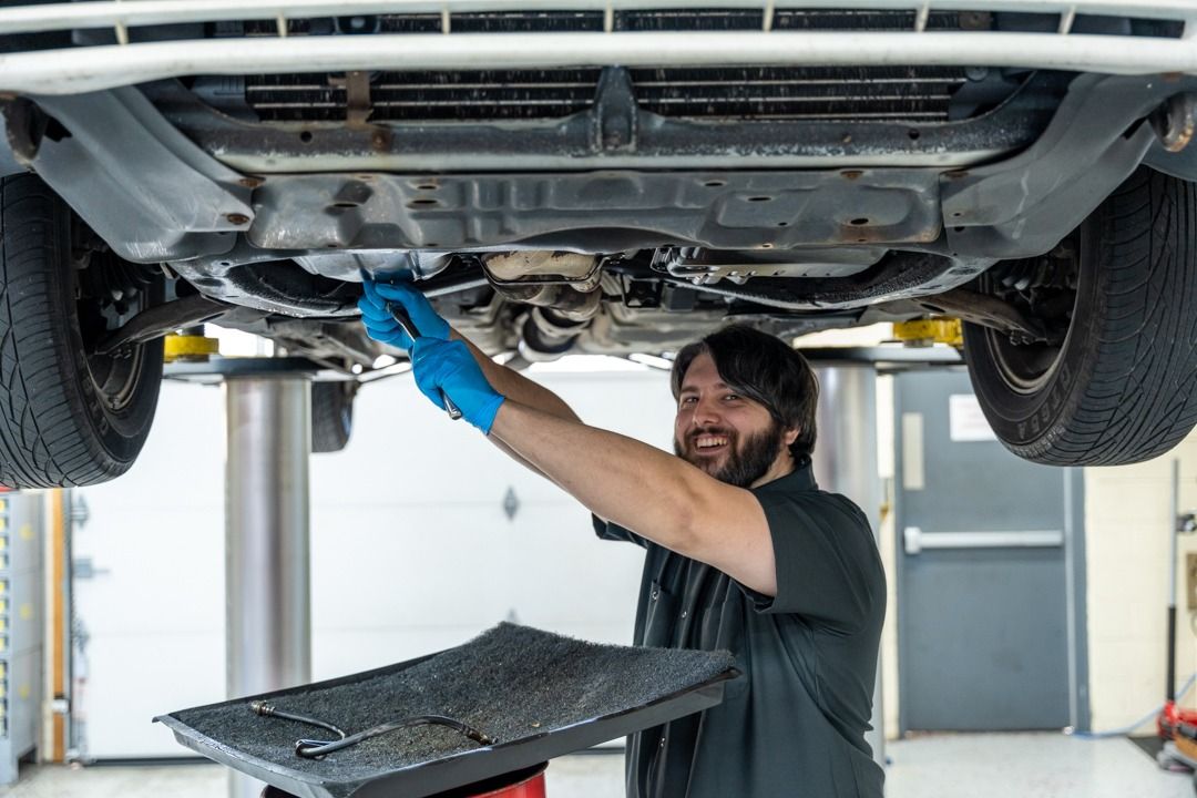 Mechanic working under a car on a lift; holding a tool; smiling; wearing gloves.