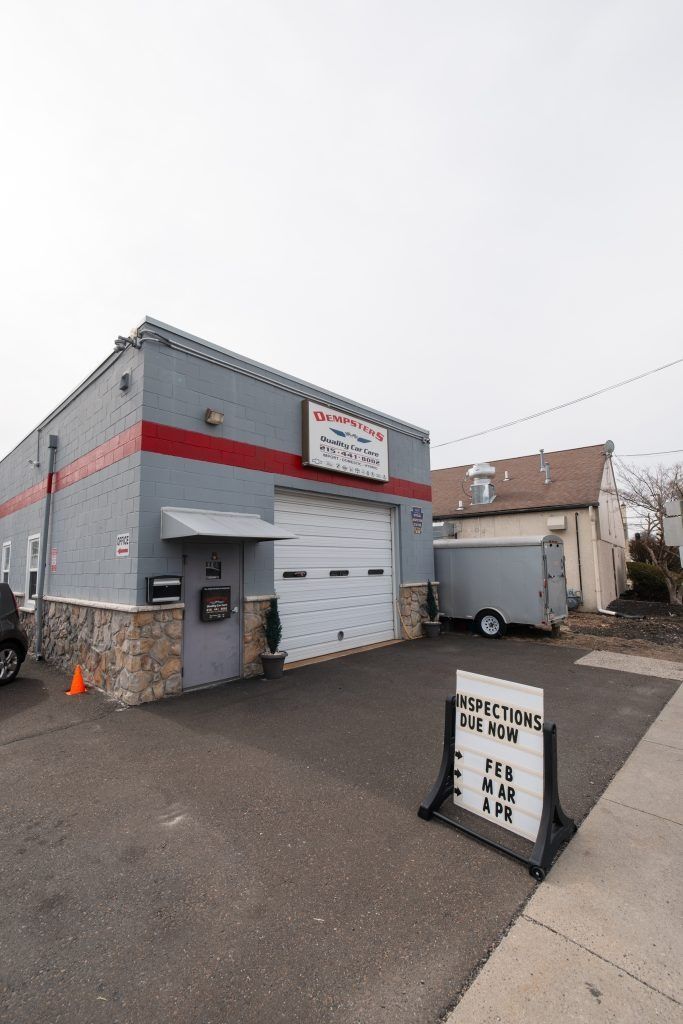 Exterior of a grey auto shop with a red stripe, a closed garage door, and a sandwich board sign on a paved lot.