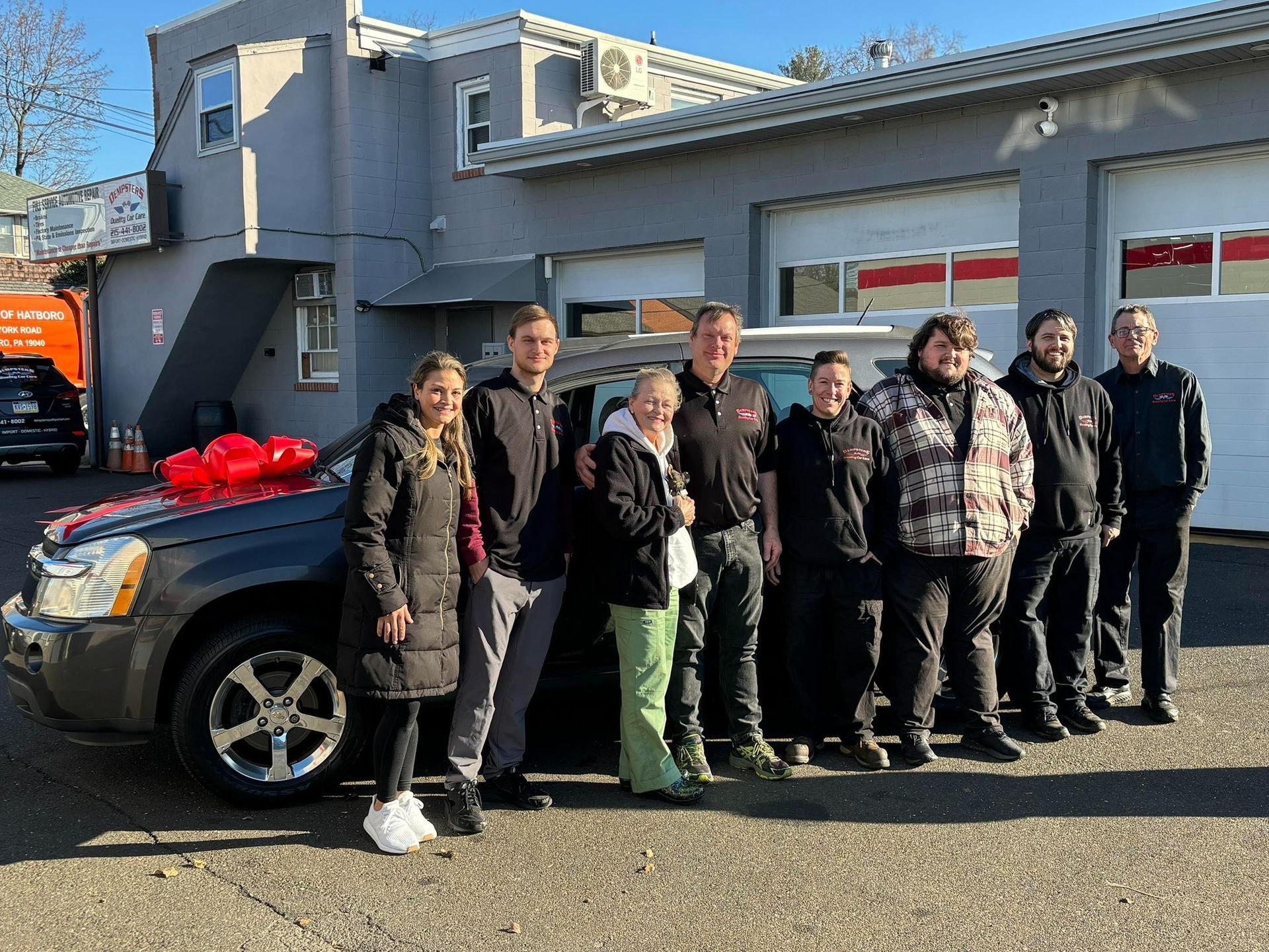 Group of people standing in front of a gray car with a red bow. Building in background.