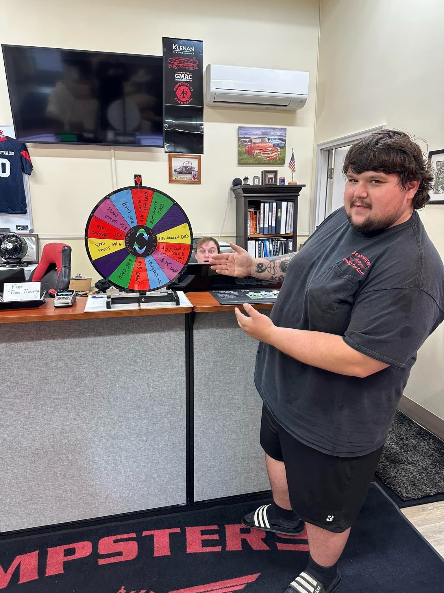 Man pointing at a prize wheel behind a counter. Another person is visible behind the wheel.