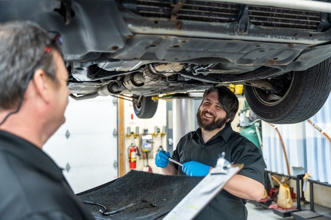 Two mechanics examine a car's underside in a garage. One holds a clipboard; another works with a wrench.