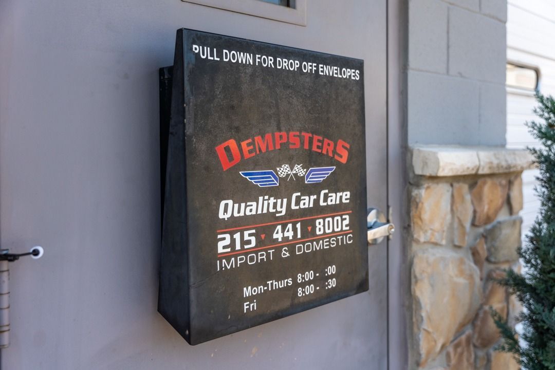 Black sign for Dempster's Quality Car Care, on a gray wall. Includes text, logo and phone number.