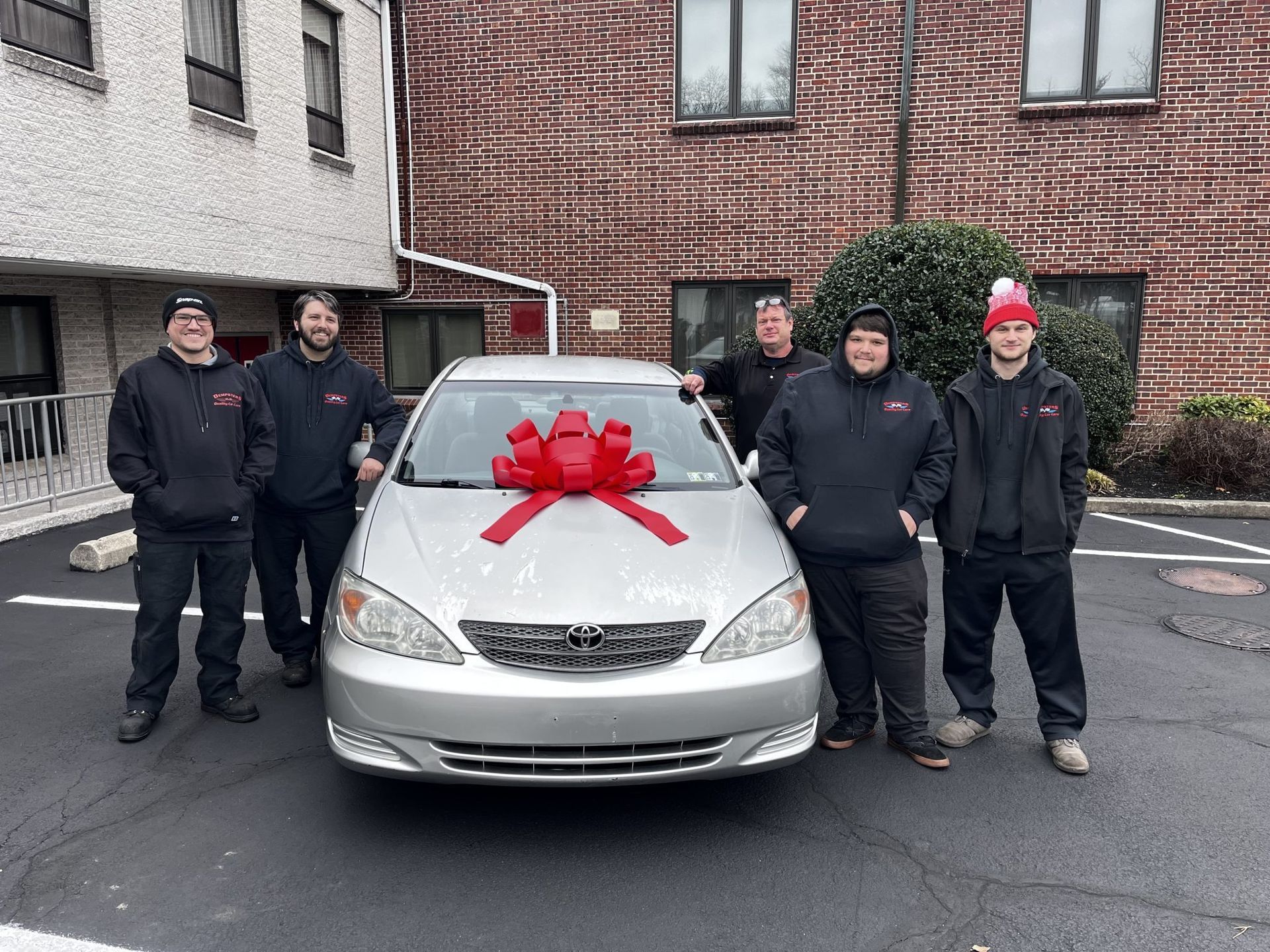 Six people stand in front of a silver car with a large red bow, in front of a brick building.