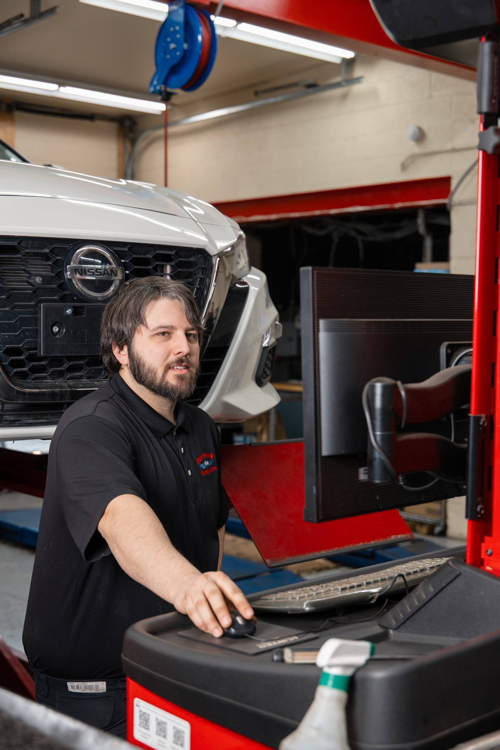 Mechanic at computer in auto shop, working on car alignment; Nissan vehicle visible.