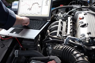 Mechanic using a laptop to diagnose a car engine. The engine is visible, along with diagnostic graphs on the laptop screen.