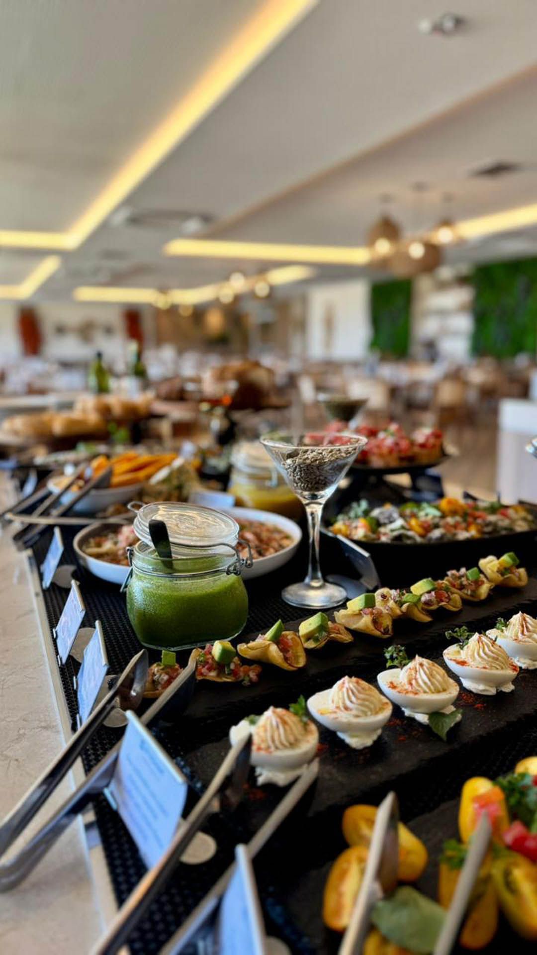 Mesa de buffet com aperitivos e sobremesas variadas em um salão de jantar de restaurante bem iluminado.