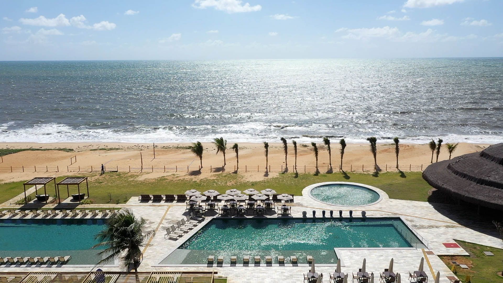 Resort à beira-mar com piscina, espreguiçadeiras, palmeiras e uma praia ensolarada.