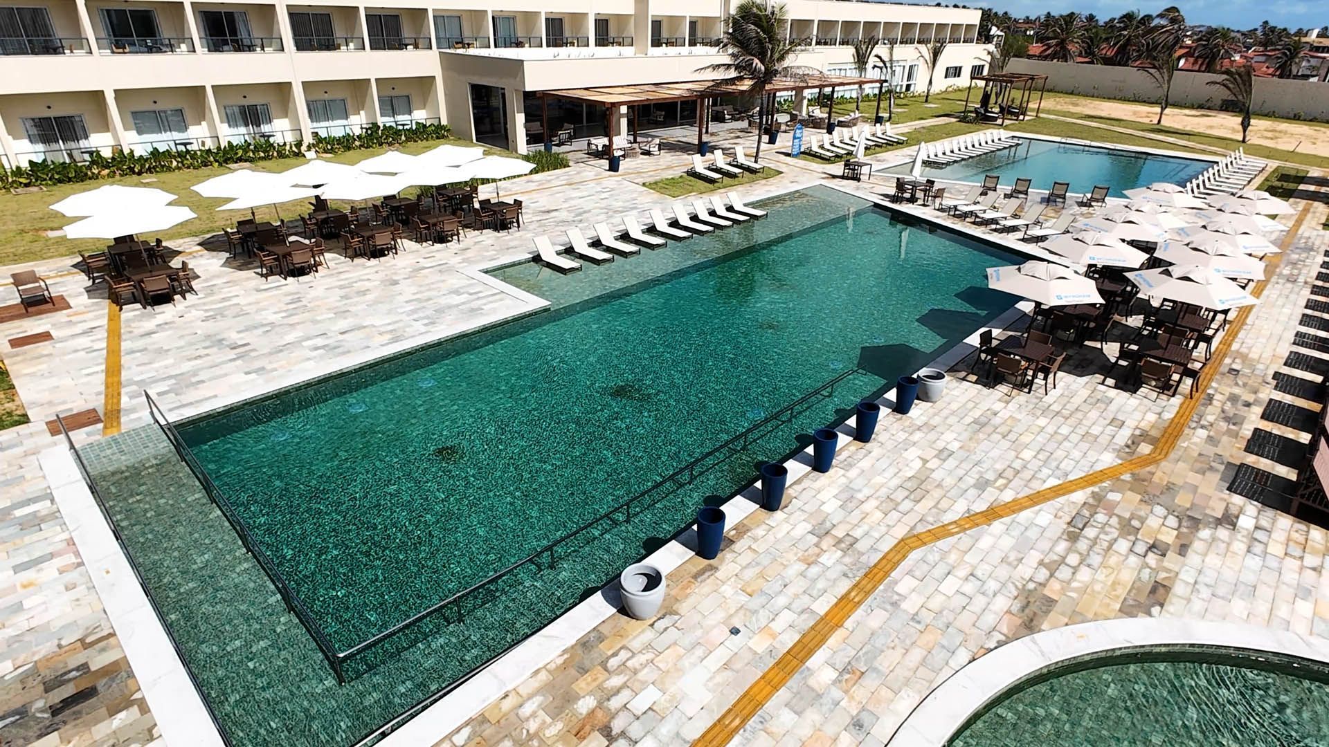 Piscina de resort com água turquesa, espreguiçadeiras e guarda-sóis brancos ao lado de um prédio de hotel.