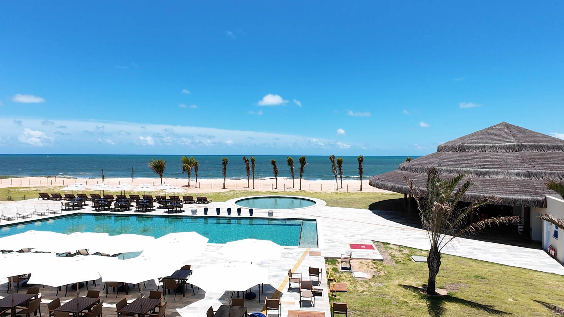 Resort à beira-mar com piscina, palmeiras, guarda-sóis brancos e um pavilhão com teto de palha junto ao oceano.