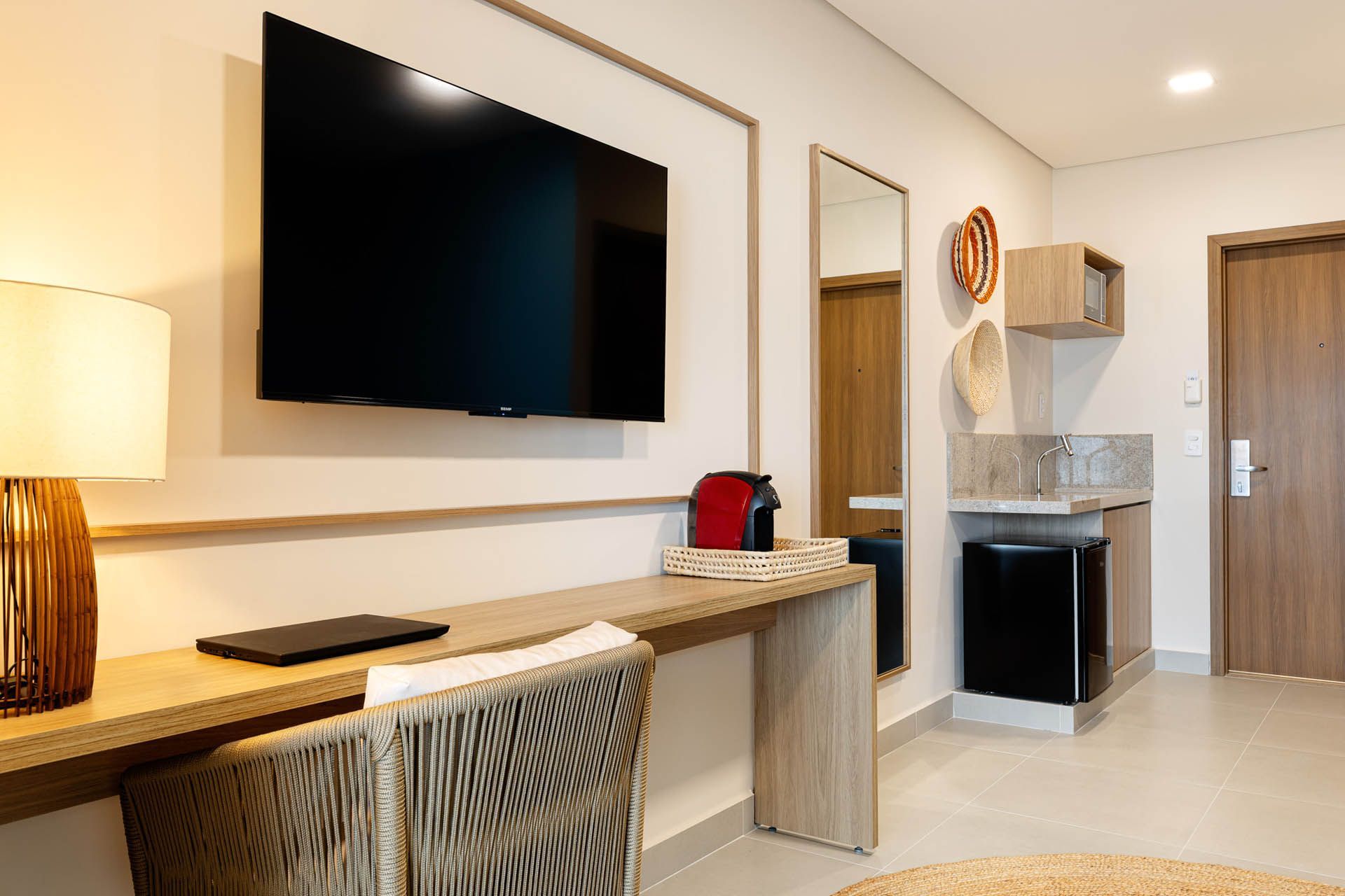 Quarto de hotel moderno com TV de parede, escrivaninha, cadeira, espelho, minicozinha e porta de entrada.