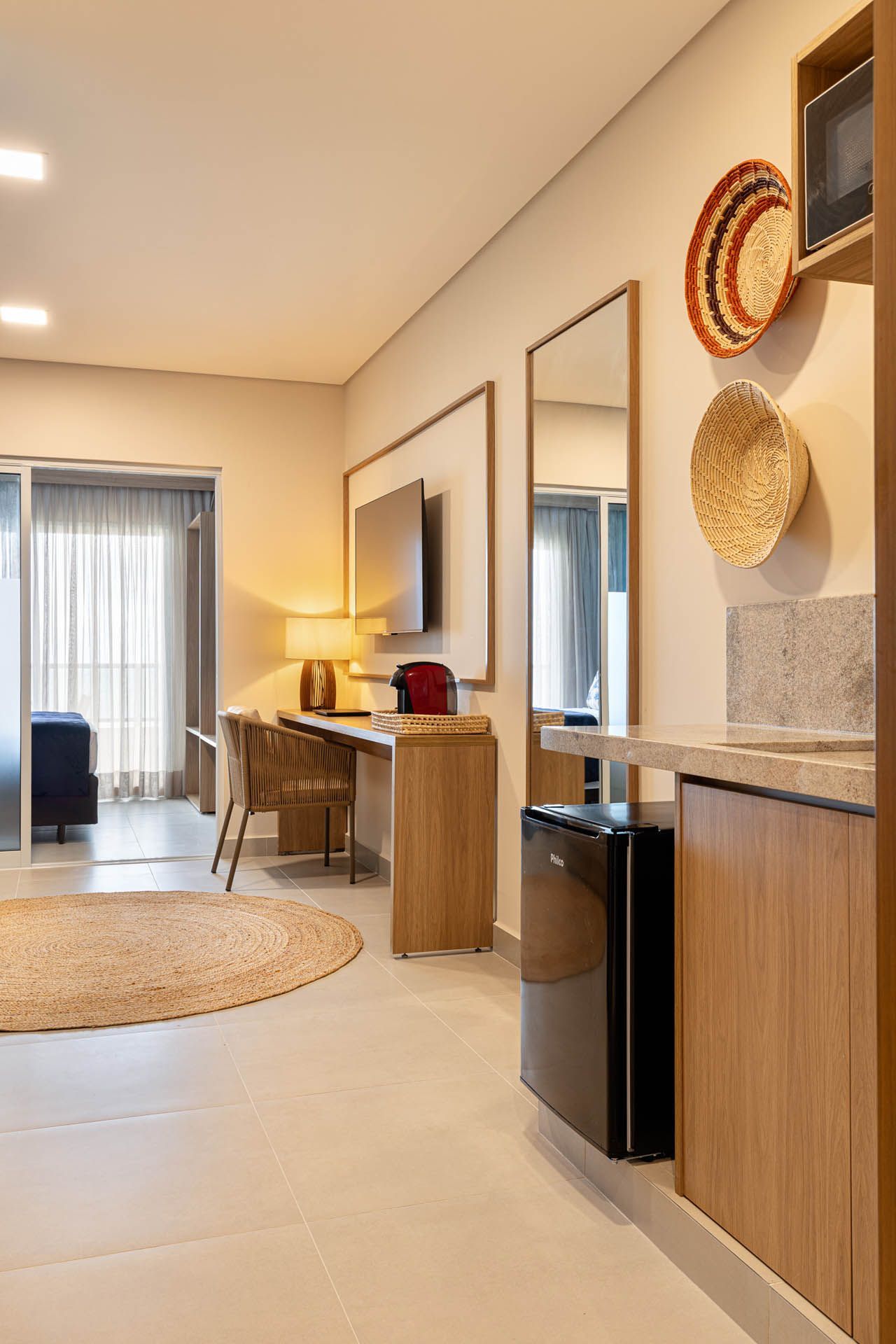 Quarto de hotel moderno com escrivaninha, espelho, luminária, tapete redondo e portas de varanda visíveis ao fundo.