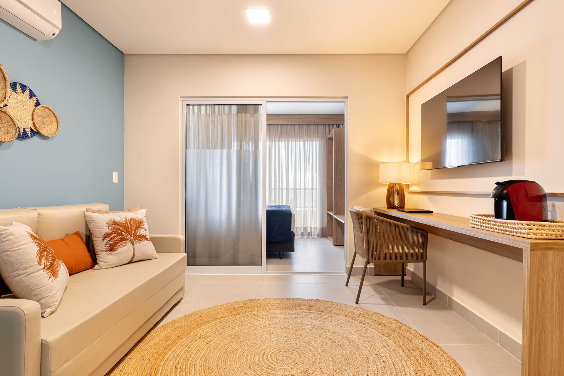 Quarto de hotel moderno com sofá, escrivaninha, TV de parede e portas de vidro deslizantes que dão para uma varanda.