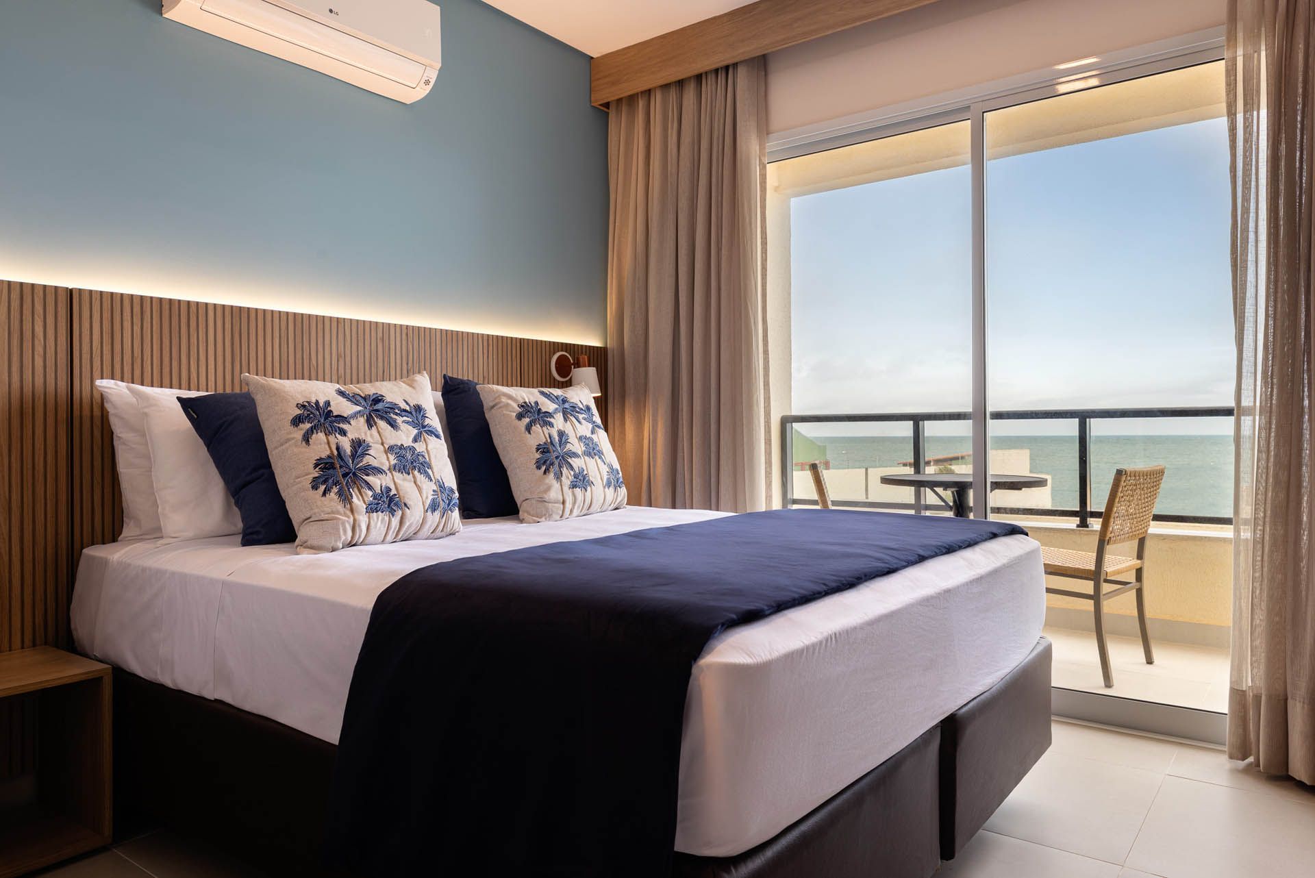 Quarto moderno com cama arrumada, detalhes em azul e portas de varanda que se abrem para uma vista ensolarada do oceano.