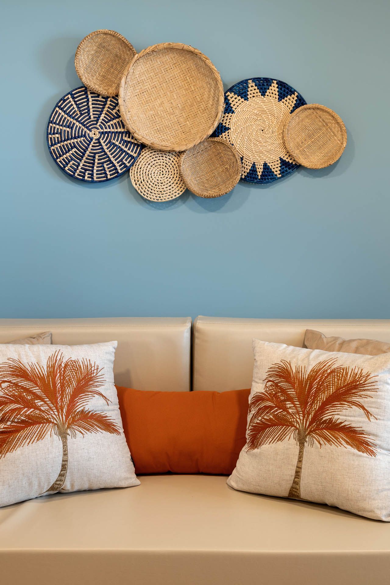 Sofá bege com almofadas laranja sob decoração de parede em tecido trançado sobre uma parede azul clara.