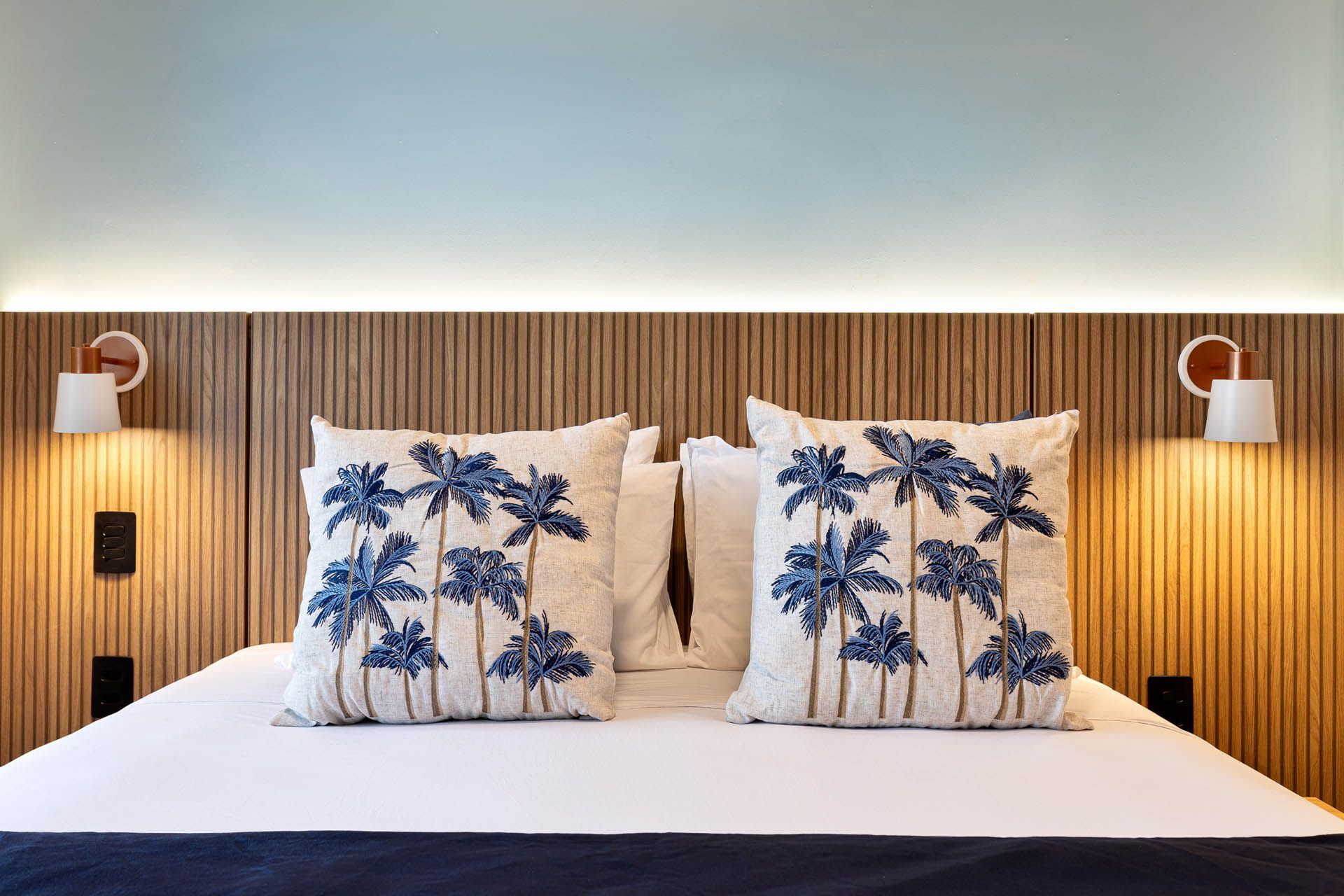 Cama impecavelmente arrumada com roupa de cama branca e almofadas com estampa de palmeiras, encostada em uma cabeceira de madeira.
